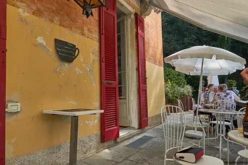 Brunate coffee shop terrace This is an architecture photograph taken in the late morning during the summer season, showing the terrace of the Brunate coffee shop near Como, Italy. The restaurant’s outdoor seating area features white metal chairs and tables beneath large white umbrellas, positioned in front of a yellow and red building with weathered walls and bright red shutters. The image captures people dining outdoors, protected by awnings and garden greenery, providing a relaxed summer atmosphere typical of Italian cafés. The photograph highlights the architectural details and ambiance of a restaurant setting in the hills above Como.